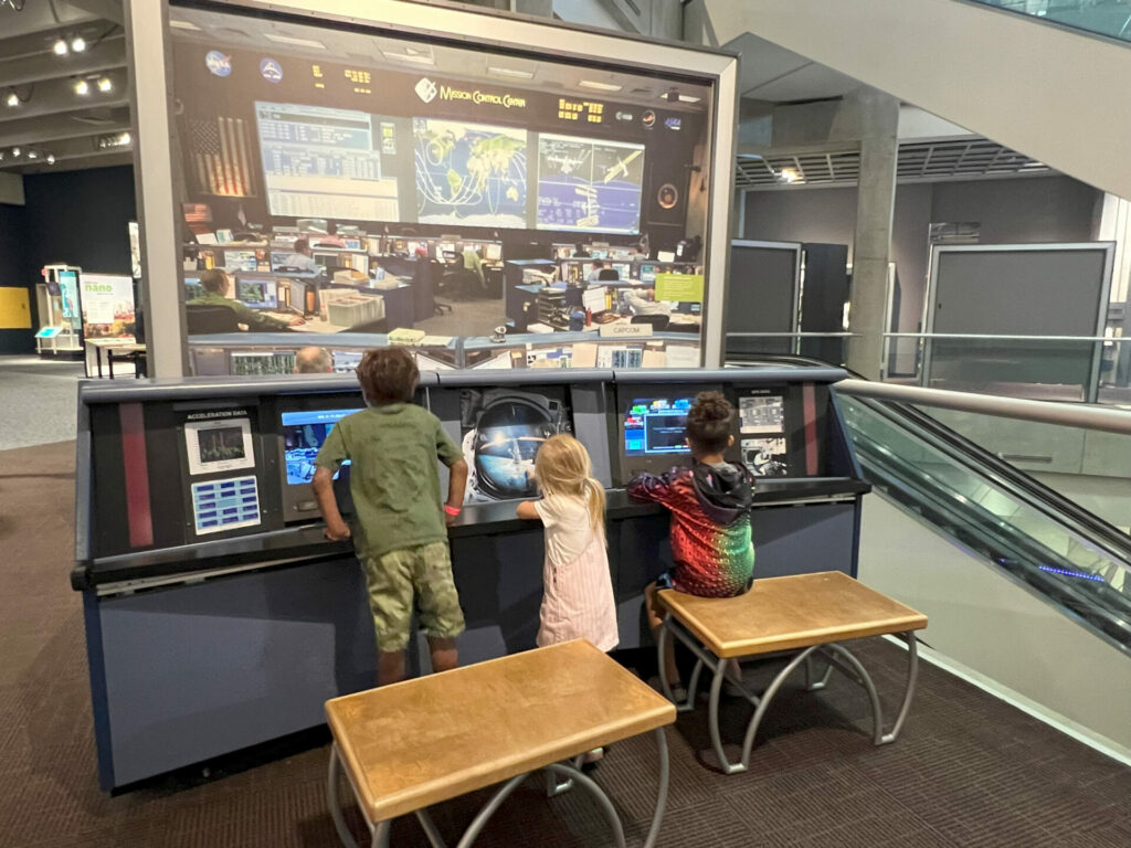 Three children exploring different science stations from being at the shuttle control center, to playing in a ball pit and learning about sounds. 