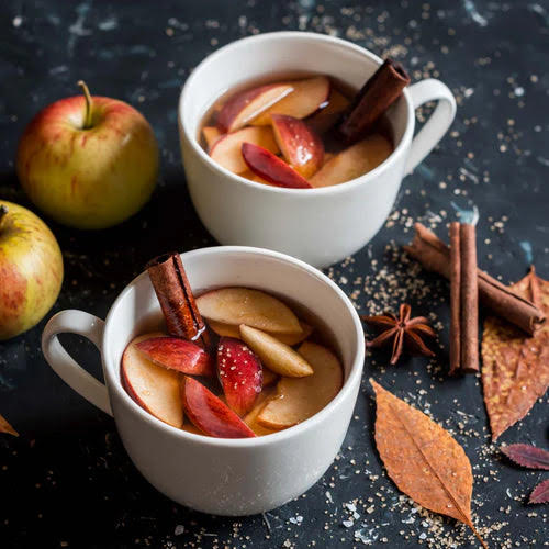Image of two white cups filled with spiced apple cider topped with sliced apples and cinnamon sticks.