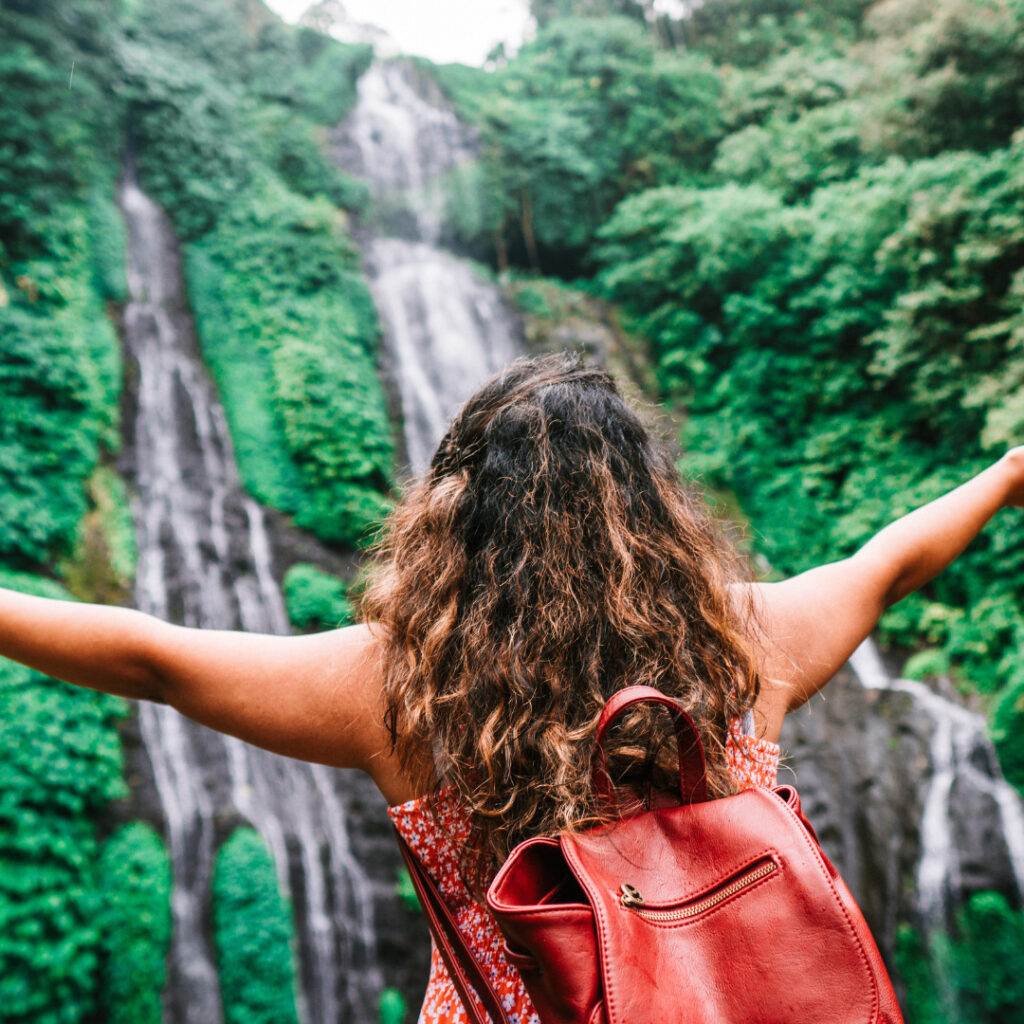 Camera angle from behind shows a young woman with a back pack and arms open wide staring up in awe at a waterfall in the jungle