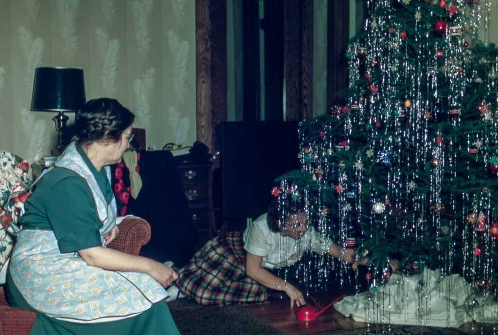 Celebrating Chiristmas memories: Circa 1960s woman on couch watching young girl putting something under a christmas tree 
decorated with lights, ornaments and silver tinsel. 