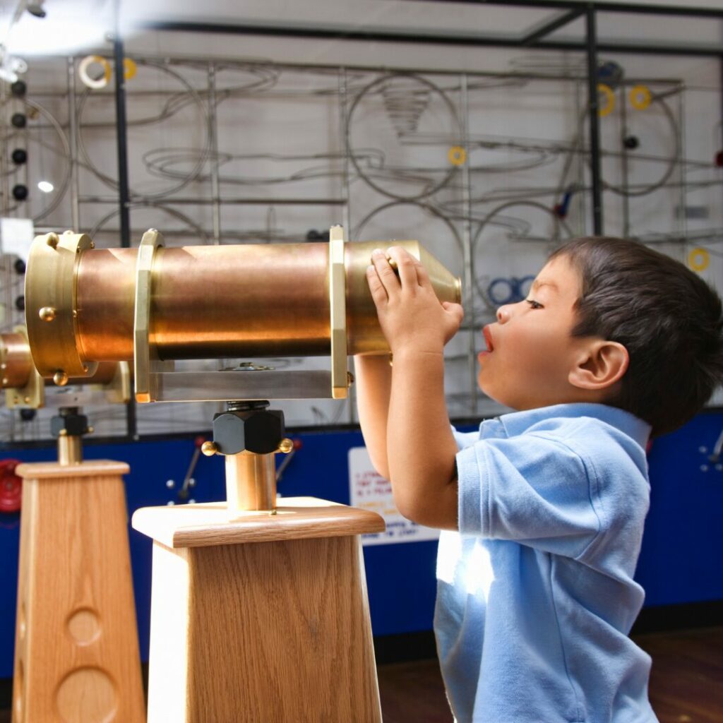 A young boy is enjoying indoor winter activites at a science center as he stretches to see out the lens of a high powered telescope.
