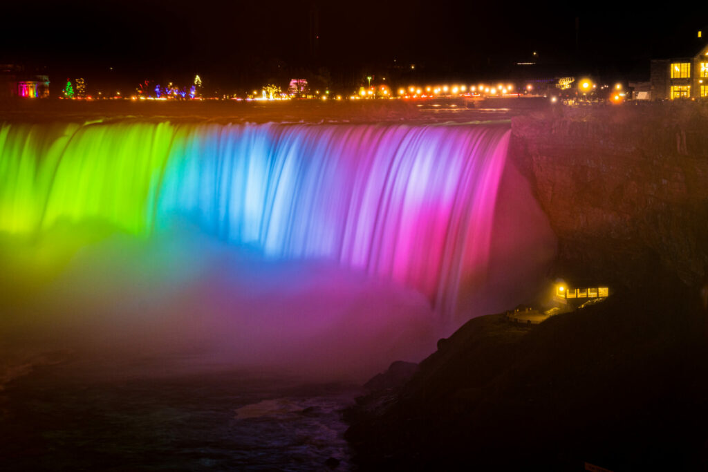 Niagara Falls at night with a rainbow of colors illuminating the falls and a city skyline in the distance