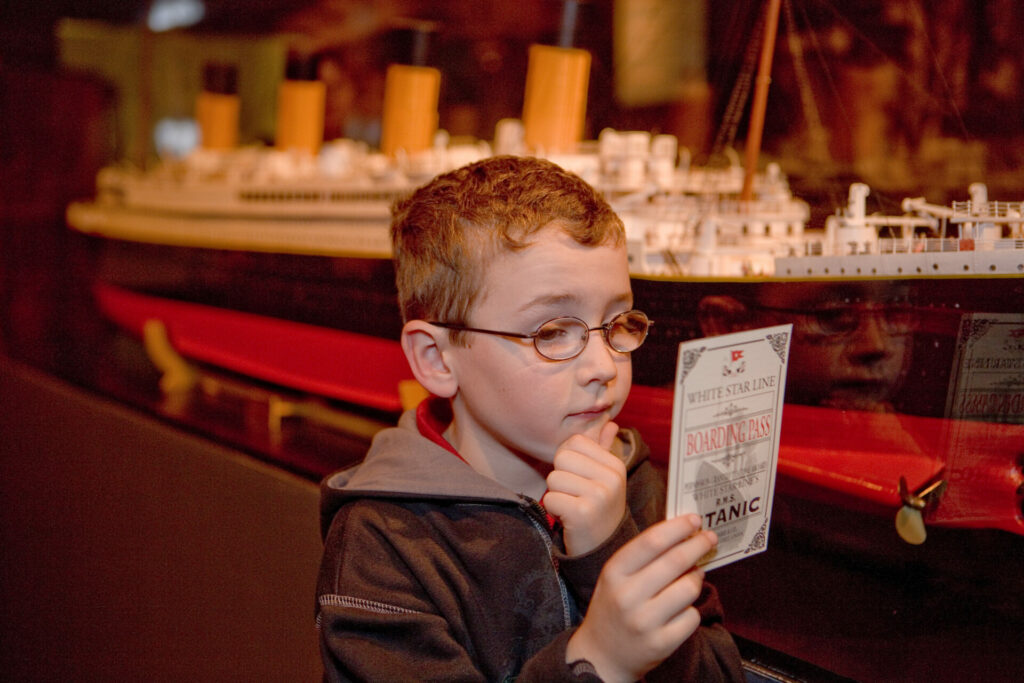 A young boy is looking at his boarding pass for the Titanic in front of a model of the ship at COSI Science Museum