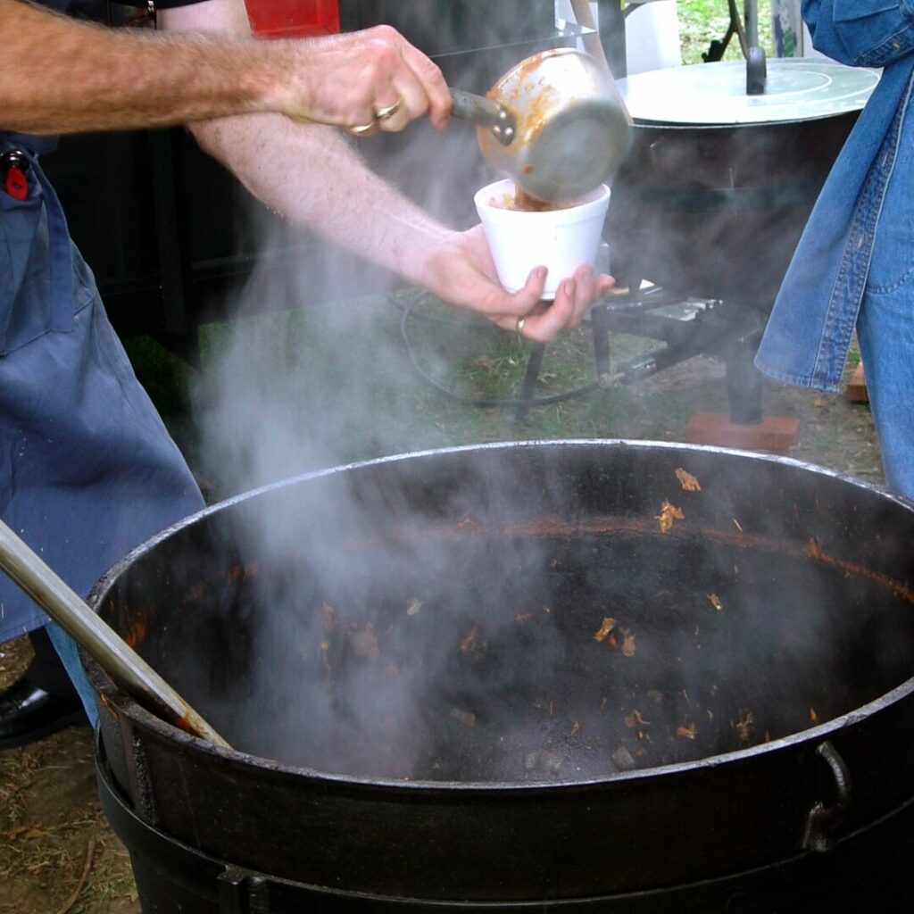 A large kettle over an open fire with hot steam emitting as people ladle out burgoo stew