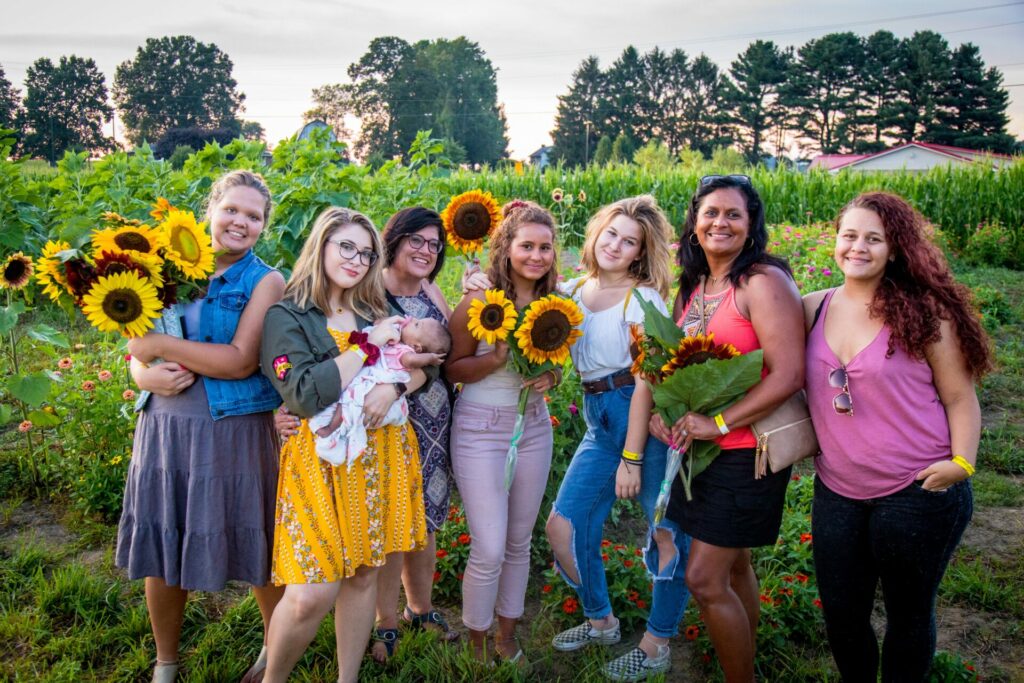 A diverse group of women some holding sunflowers pose in front of a field of sunflowers at the Coshocton Sunflower festival