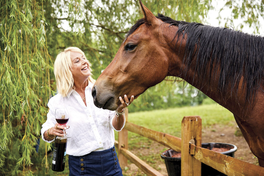 A woman holding a glass of wine as she pets a brown horse under the chin